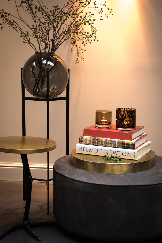 Glass ball vase on a three-legged stand, next to a chicken-legged side table, and a pair of votives atop a stack of books.